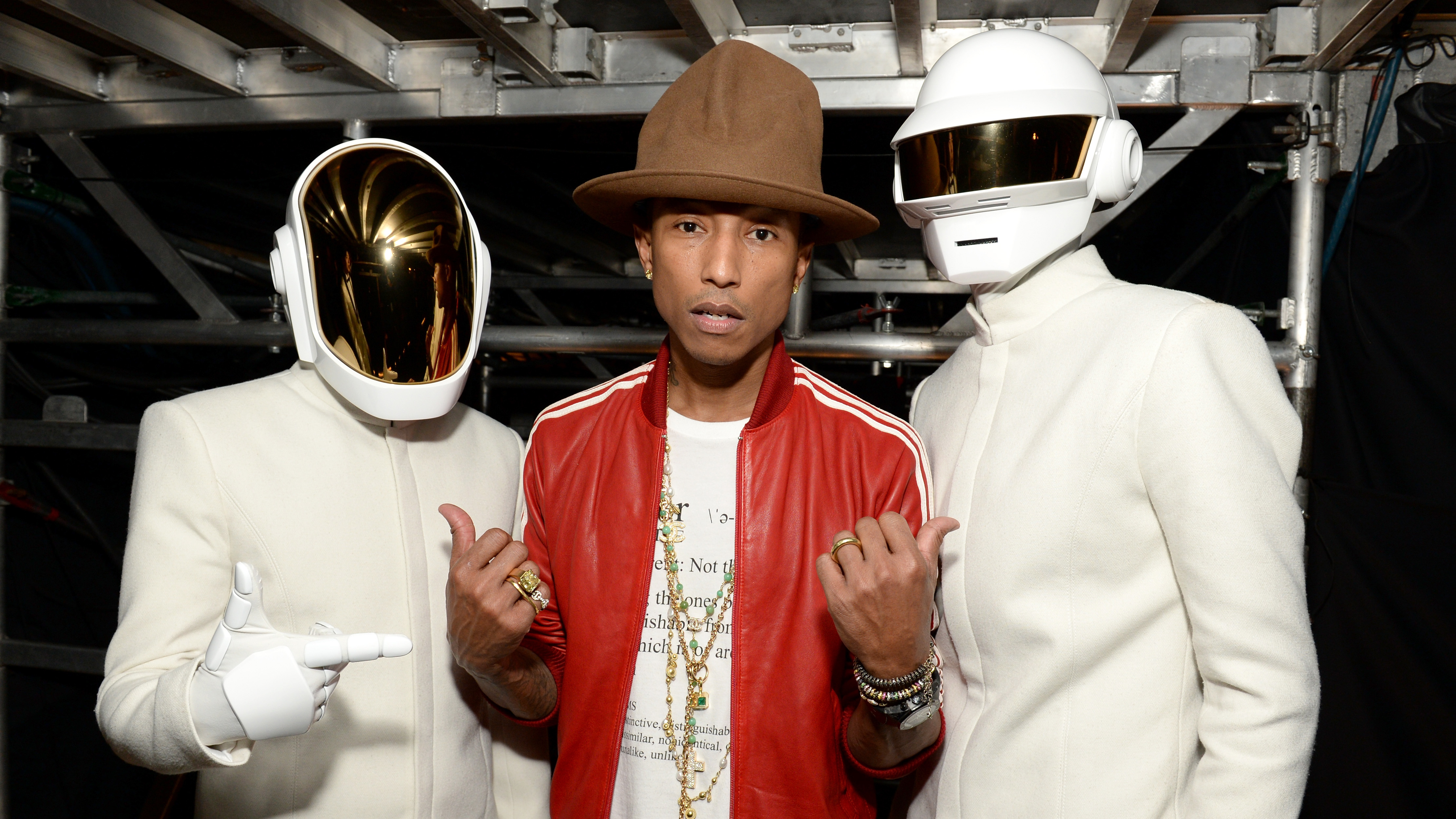 Pharrell Williams in a brown hat and red jacket poses with Daft Punk, wearing white suits and helmets.